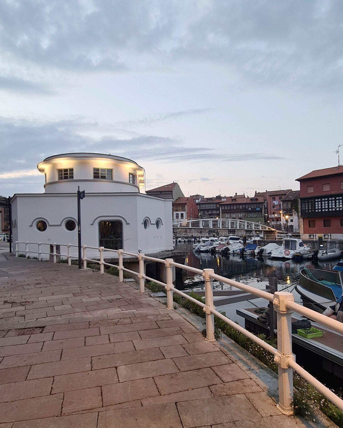 En la costa de Llanes se puede ver un edificio blanco moderno con ventanas redondas, rodeado por una barandilla. En el puerto deportivo adyacente hay varios barcos atracados, mientras que los edificios tradicionales se alzan bajo un cielo nublado de verano.