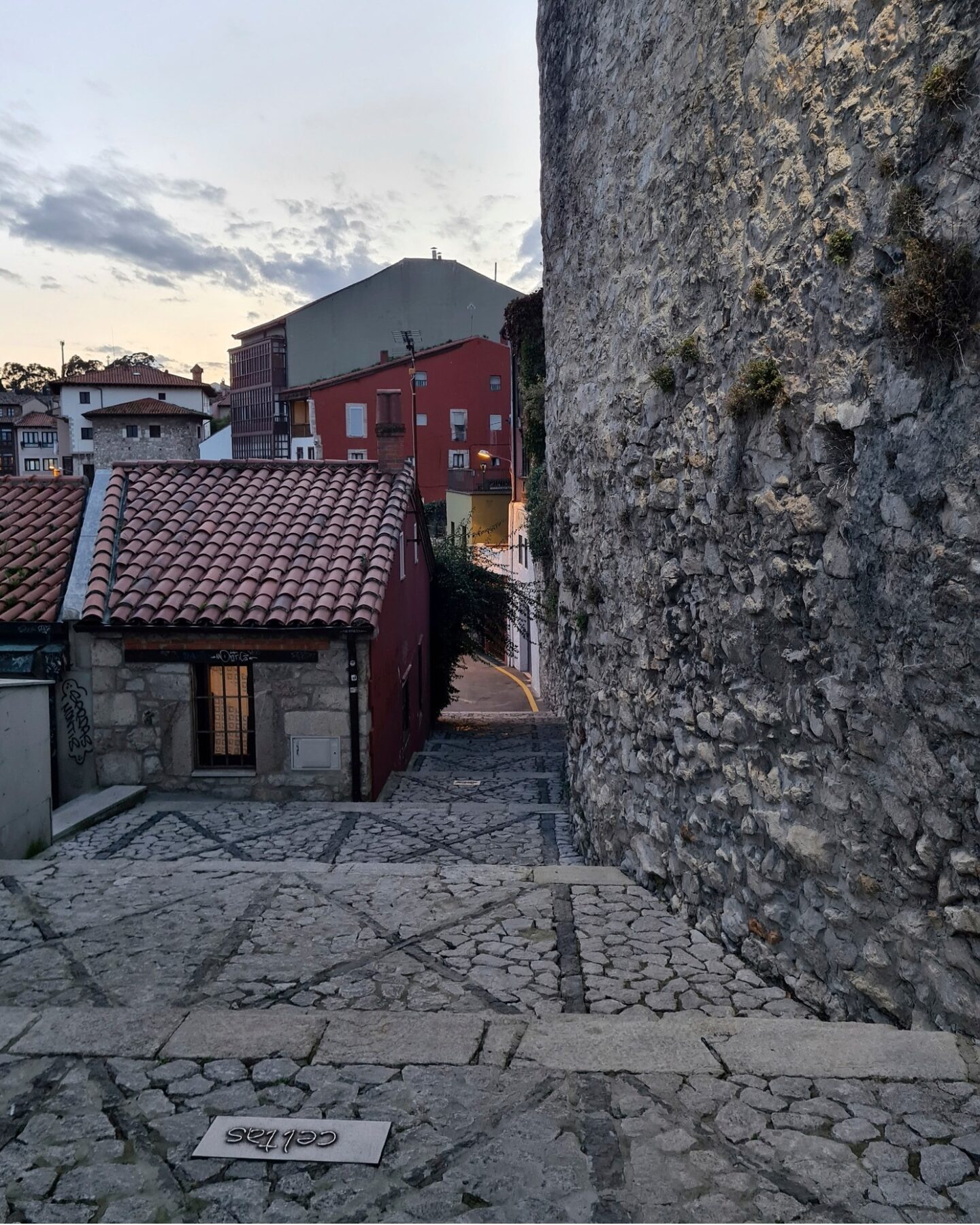 Un estrecho camino adoquinado conduce entre paredes de piedra texturizada y encantadores edificios residenciales con techos de tejas rojas. Mientras la música se desliza suavemente por el aire de la tarde, el cielo está parcialmente nublado, lo que agrega un toque sereno a esta encantadora escena.