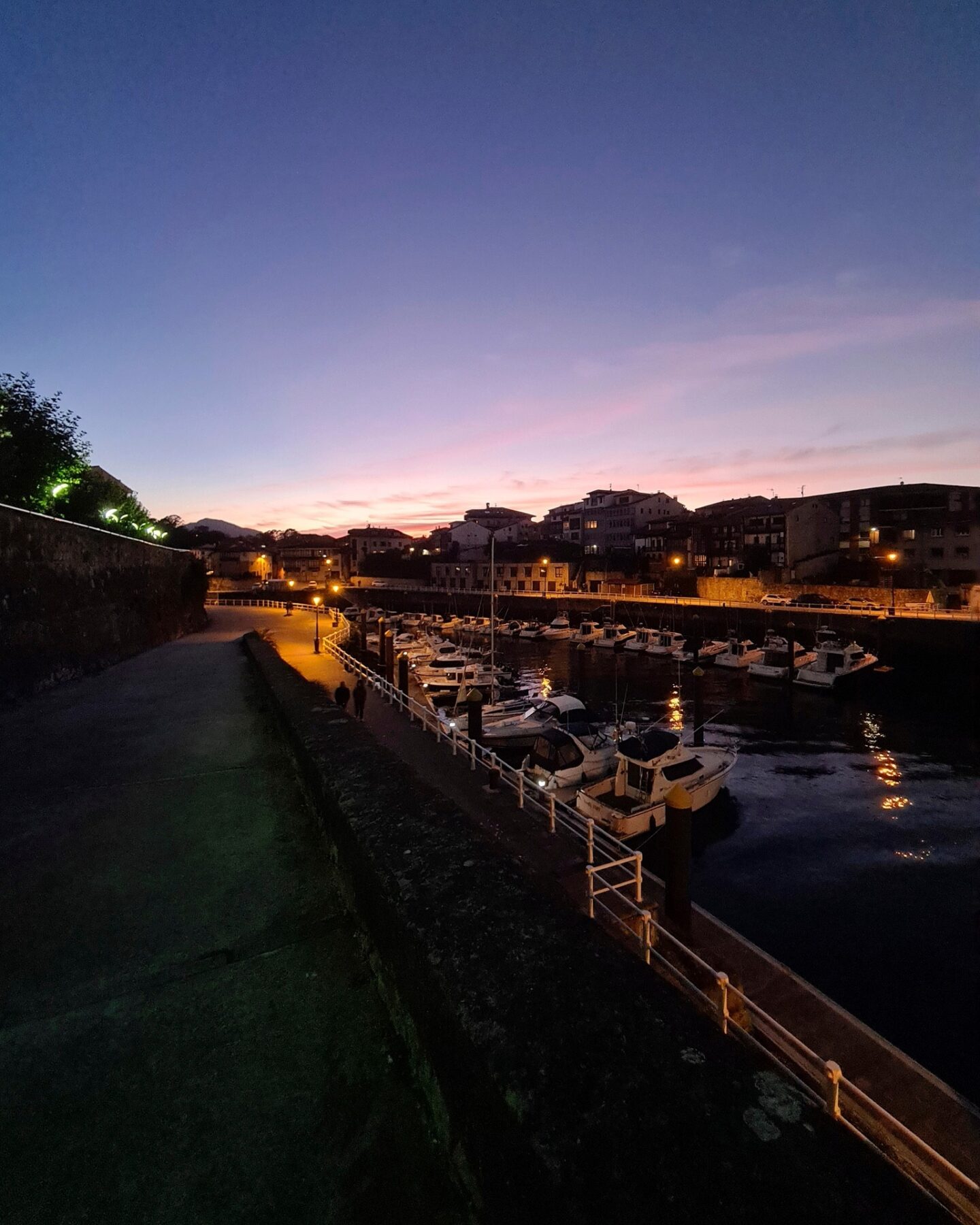 un puerto deportivo al anochecer con varios barcos atracados en el agua evoca la sensación de nos extrañamos. las farolas iluminan el paseo a la izquierda, mientras que los edificios y las colinas se ven al fondo contra un cielo rosa y violeta que se desvanece.