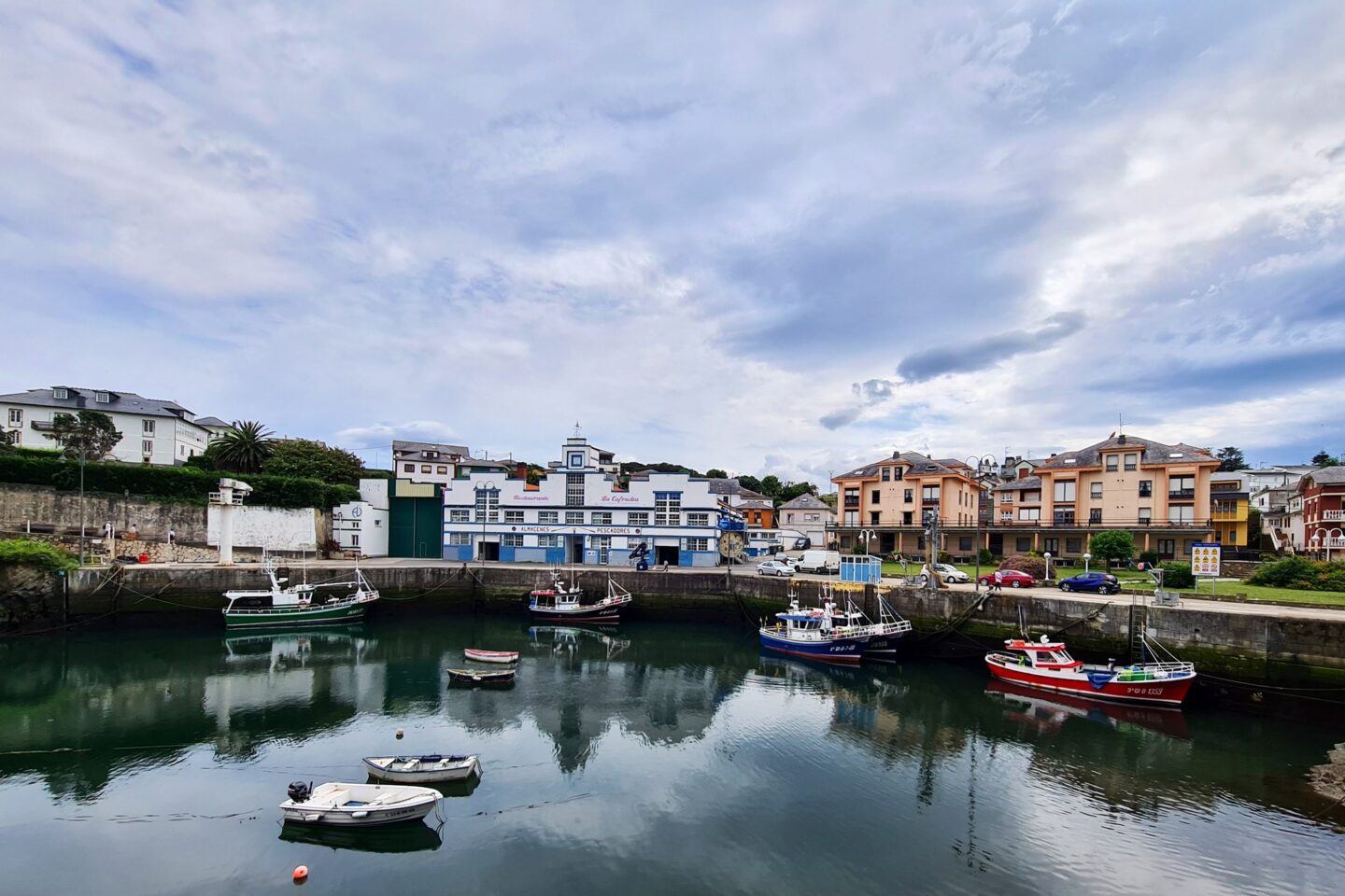 un pequeño puerto con varios barcos atracados en las tranquilas aguas de puerto de vega, parte del pintoresco mar cantábrico. al fondo, se alzan edificios coloridos bajo un cielo nublado y, cerca, encantadores restaurantes de mariscos esperan con delicias de marisco fresco.