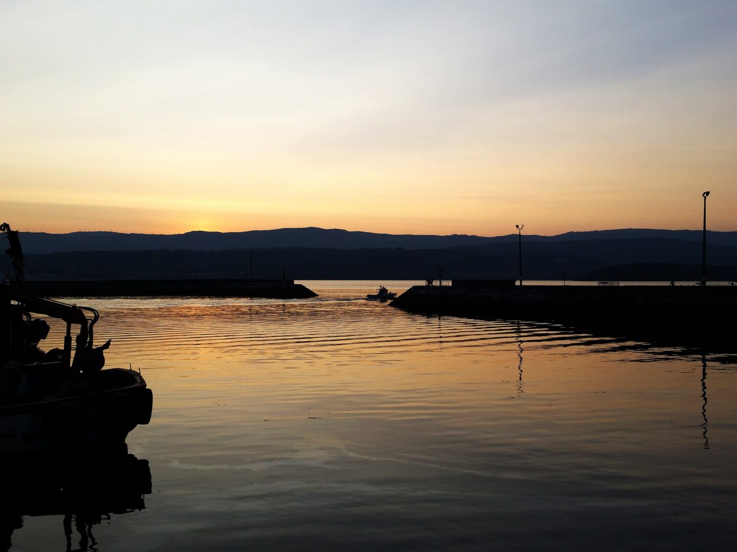 una puesta de sol serena sobre un cuerpo de agua en calma con la silueta de un pequeño barco en el centro. el cielo presenta un degradado de naranja a gris y se ven montañas al fondo. el agua refleja los colores del cielo.