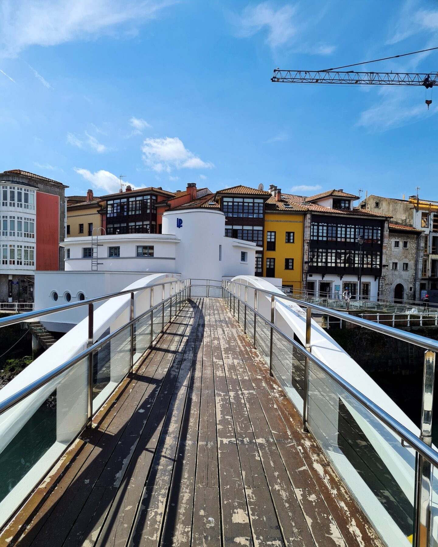 un puente de madera con barandillas de vidrio conduce sobre un cuerpo de agua hacia una hilera de edificios coloridos que recuerdan a otras vidas. los edificios están pintados de blanco, amarillo y rojo con grandes ventanales. se ve una grúa al fondo contra un cielo azul.